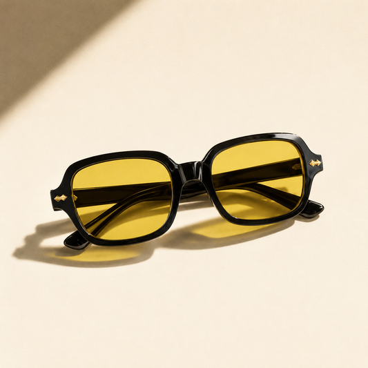 Oversized square black sunglasses with warm golden amber tinted lenses resting on a sunlit beige stone surface
