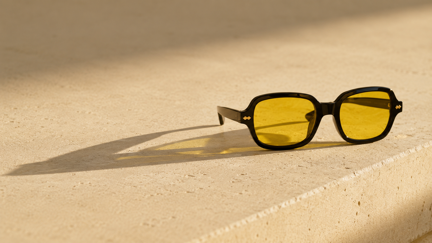 Black square sunglasses with yellow tinted lenses resting on a beige stone surface in warm sunlight.
